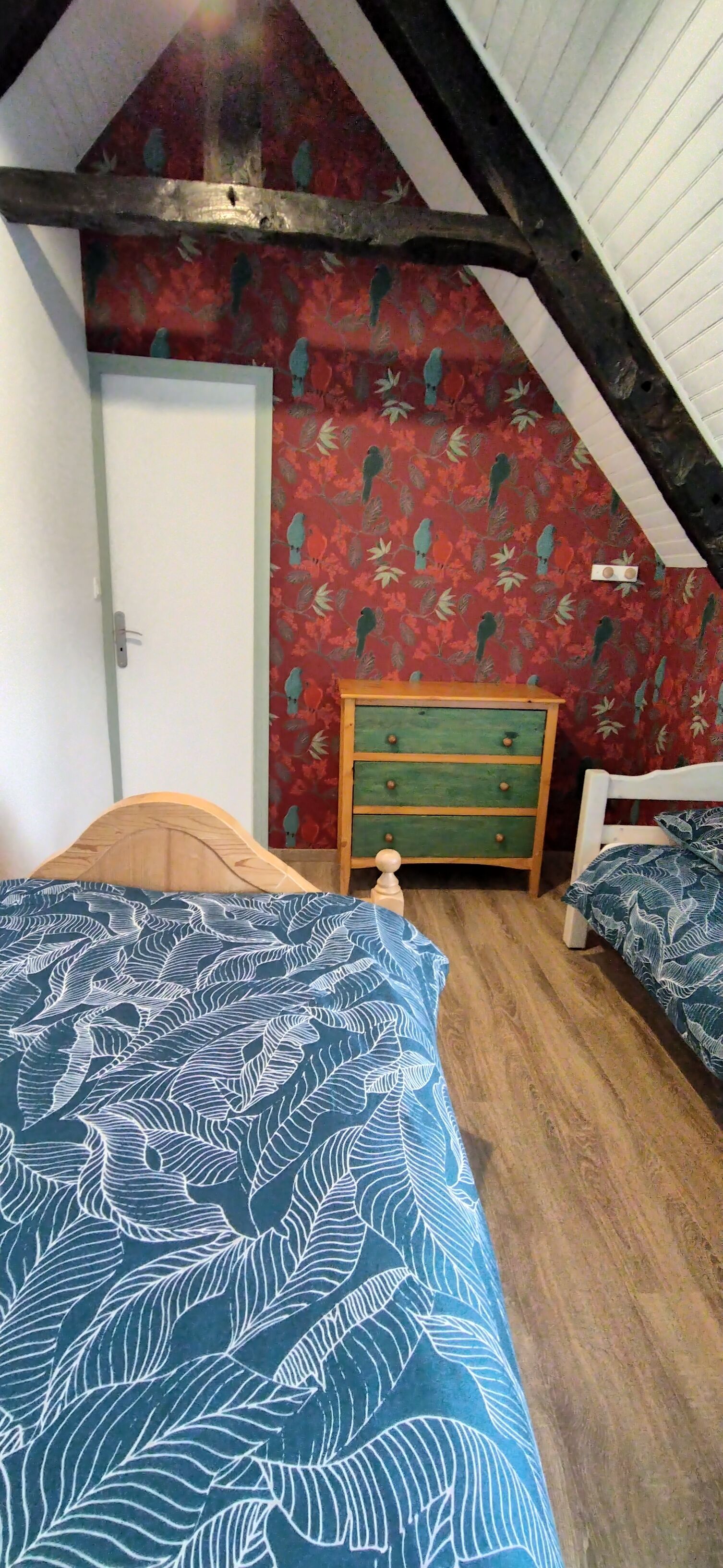 Een slaapkamer met een schuine kap, een houten bed, een groene dressoir en behang met rode bloemen.