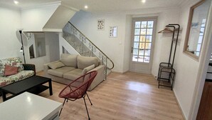 Un salon comprenant un canapé, un fauteuil, une table basse, un escalier et une porte donnant sur l’extérieur.
