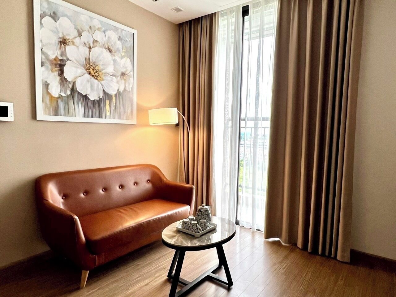A room with a brown leather sofa, a round coffee table with a tray, a floor lamp, and a large floral painting on the wall.