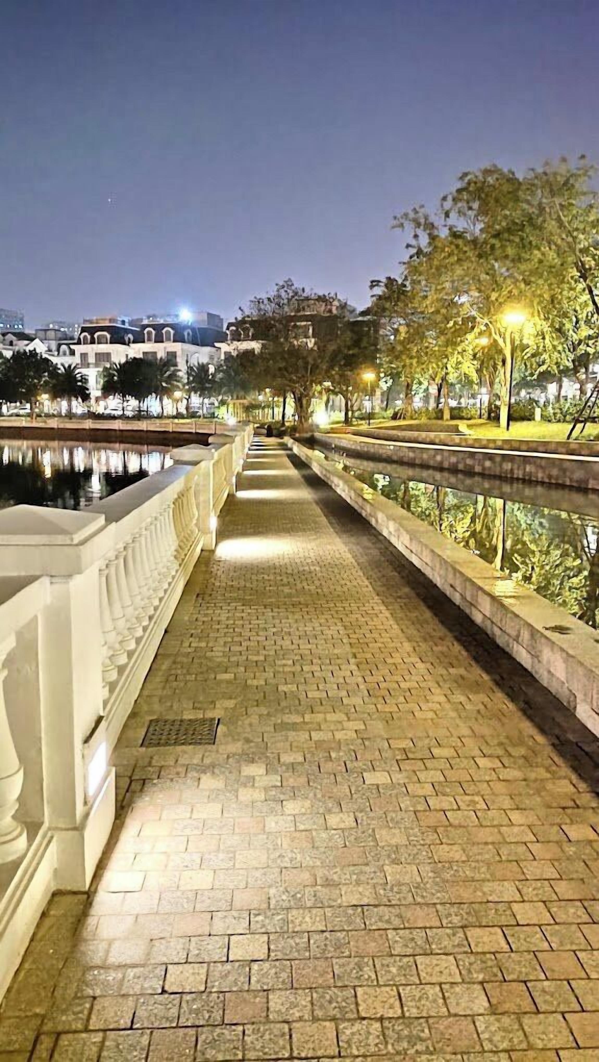 Une promenade nocturne bordée d’un côté par une étendue d’eau réfléchissante, d’un autre par un chemin pavé de briques et des arbres surmontés de lampadaires.