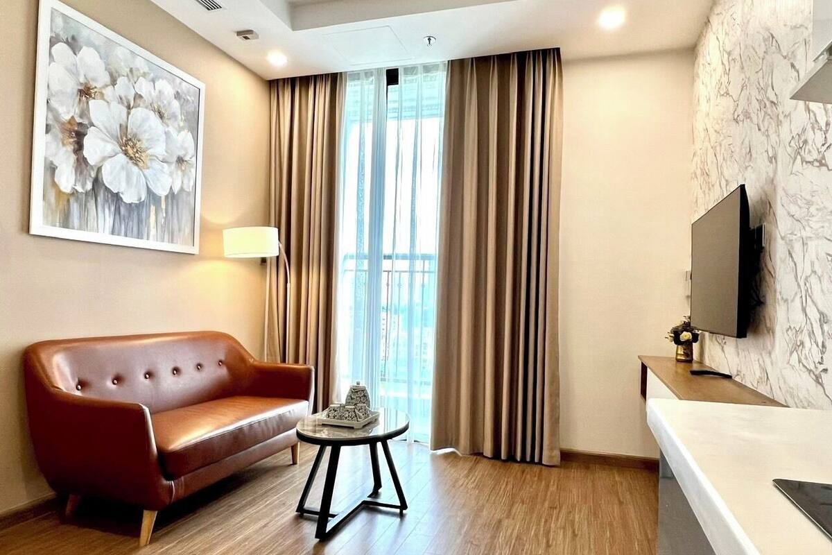 Une chambre d’hôtel moderne avec un canapé en cuir, une petite table ronde, un téléviseur fixé au mur et une grande fenêtre avec des rideaux.
