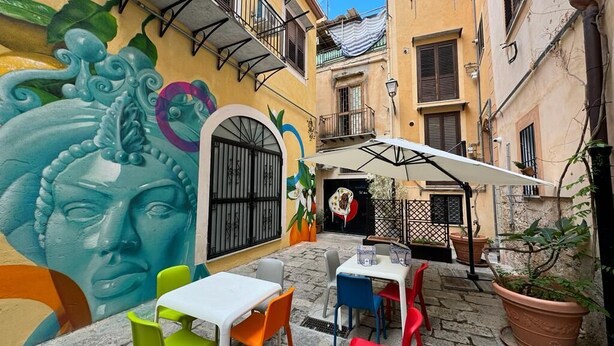 Un café al aire libre con sillas y mesas de colores, un mural en la pared y un área con pavimento de piedra.