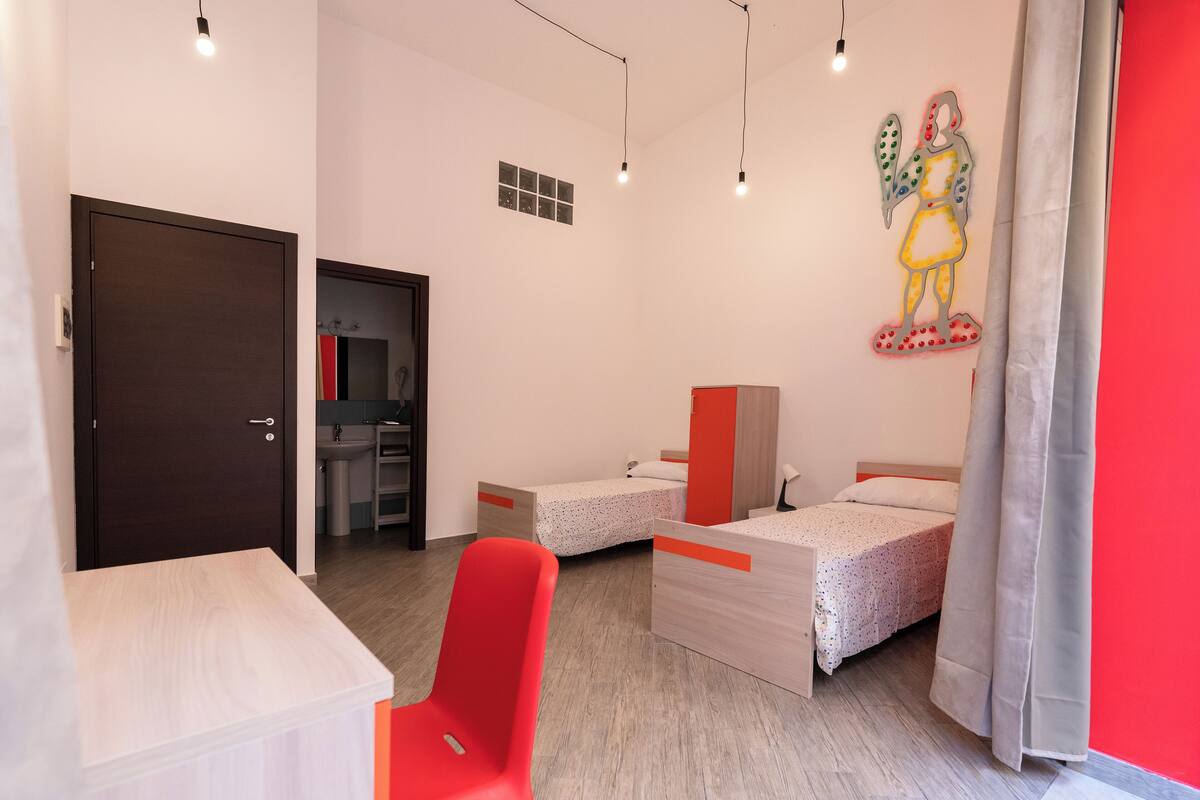 Un dormitorio moderno con dos camas individuales, un escritorio, una silla y un baño visible al fondo.