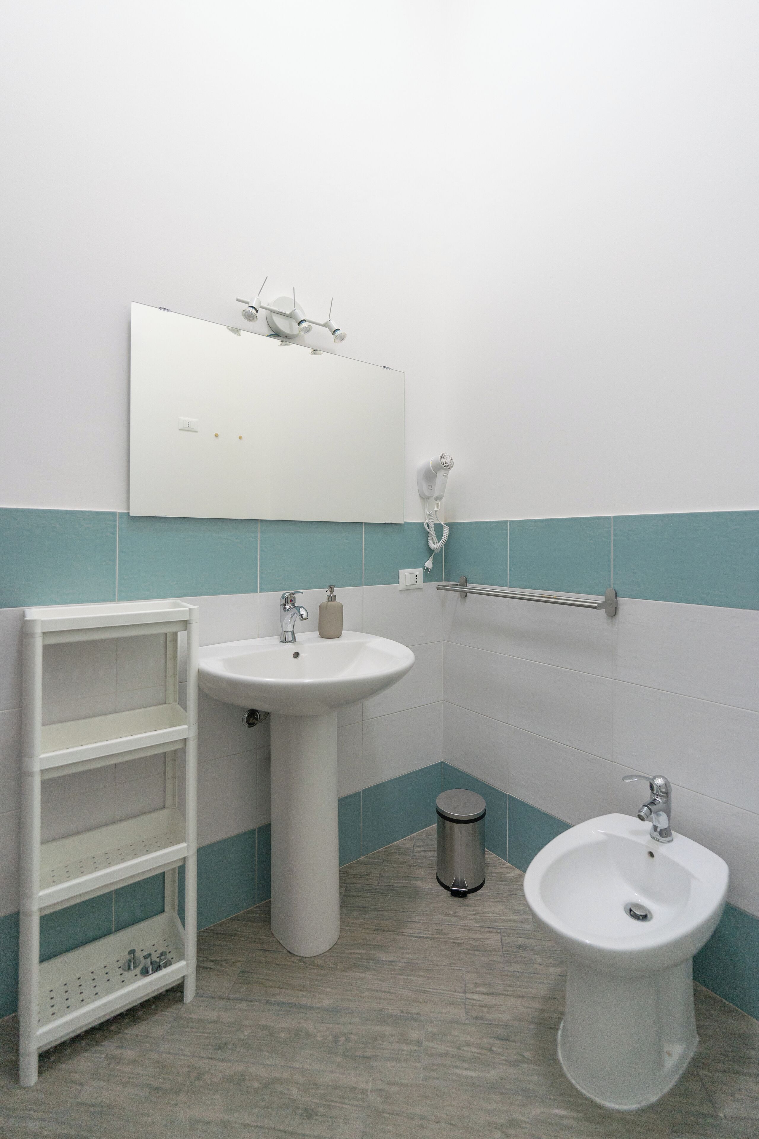 A bathroom with a white sink, a bidet, a mirror, and a shelf unit.