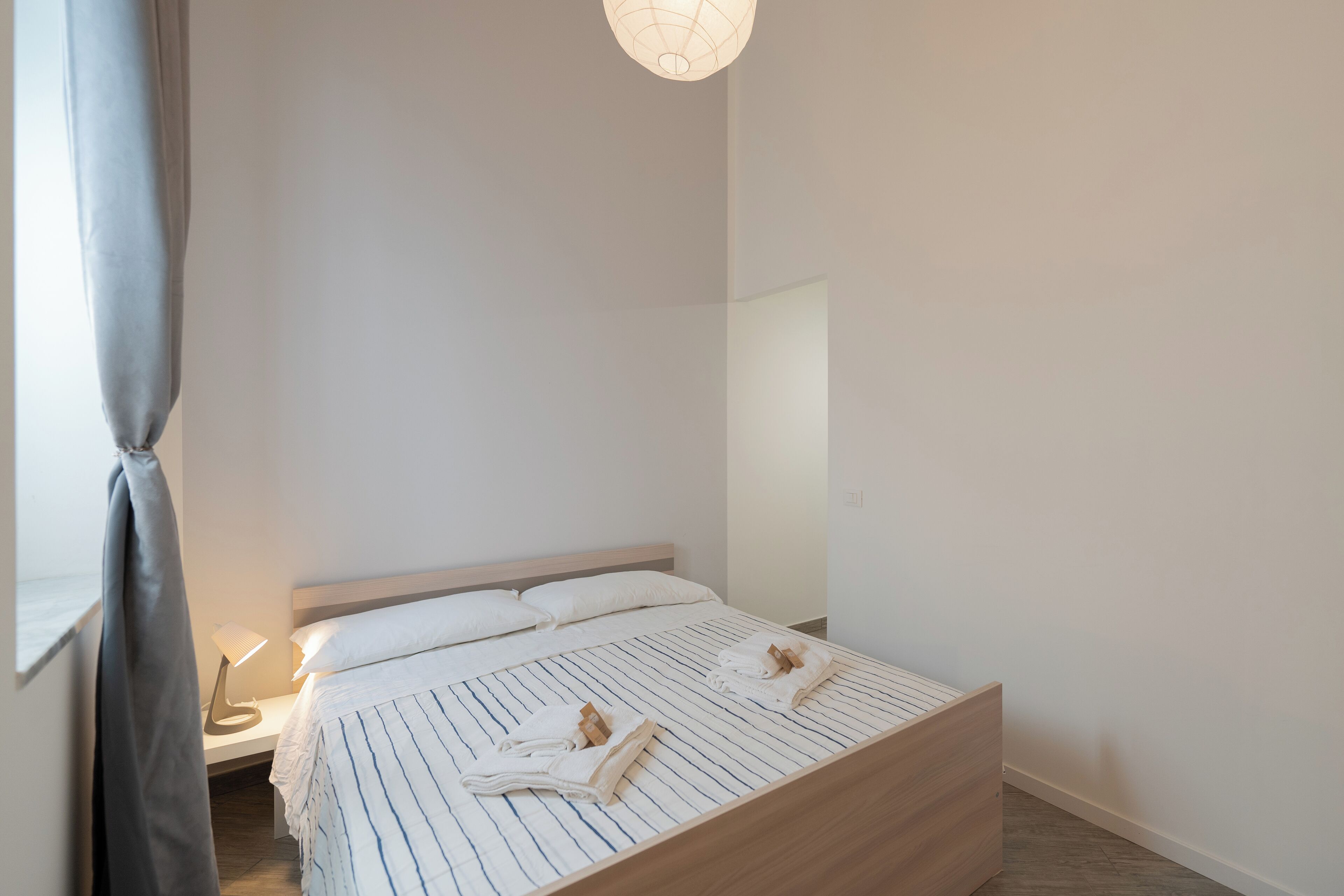 Una cama bien hecha con ropa de cama a rayas y toallas, una lámpara de noche y una cortina colgante.