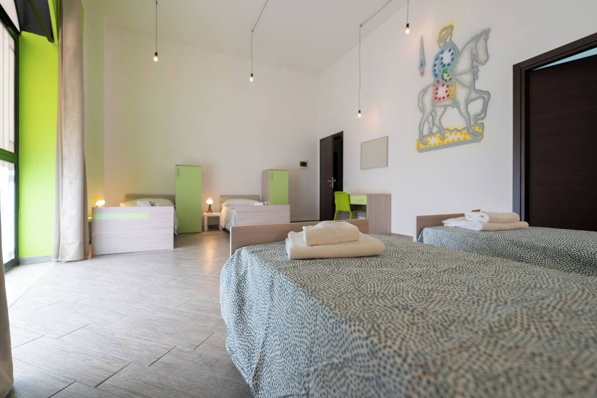 Una habitación de hotel moderna con dos camas, un ventanal grande, una pared verde y un cuadro de un caballero a caballo.