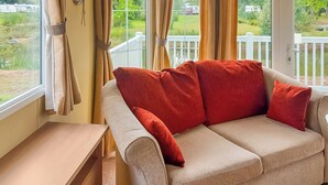 Un canapé beige agrémenté de coussins rouges, une petite table d’appoint en bois et, par la fenêtre, une vue sur une clôture blanche et la verdure environnante.