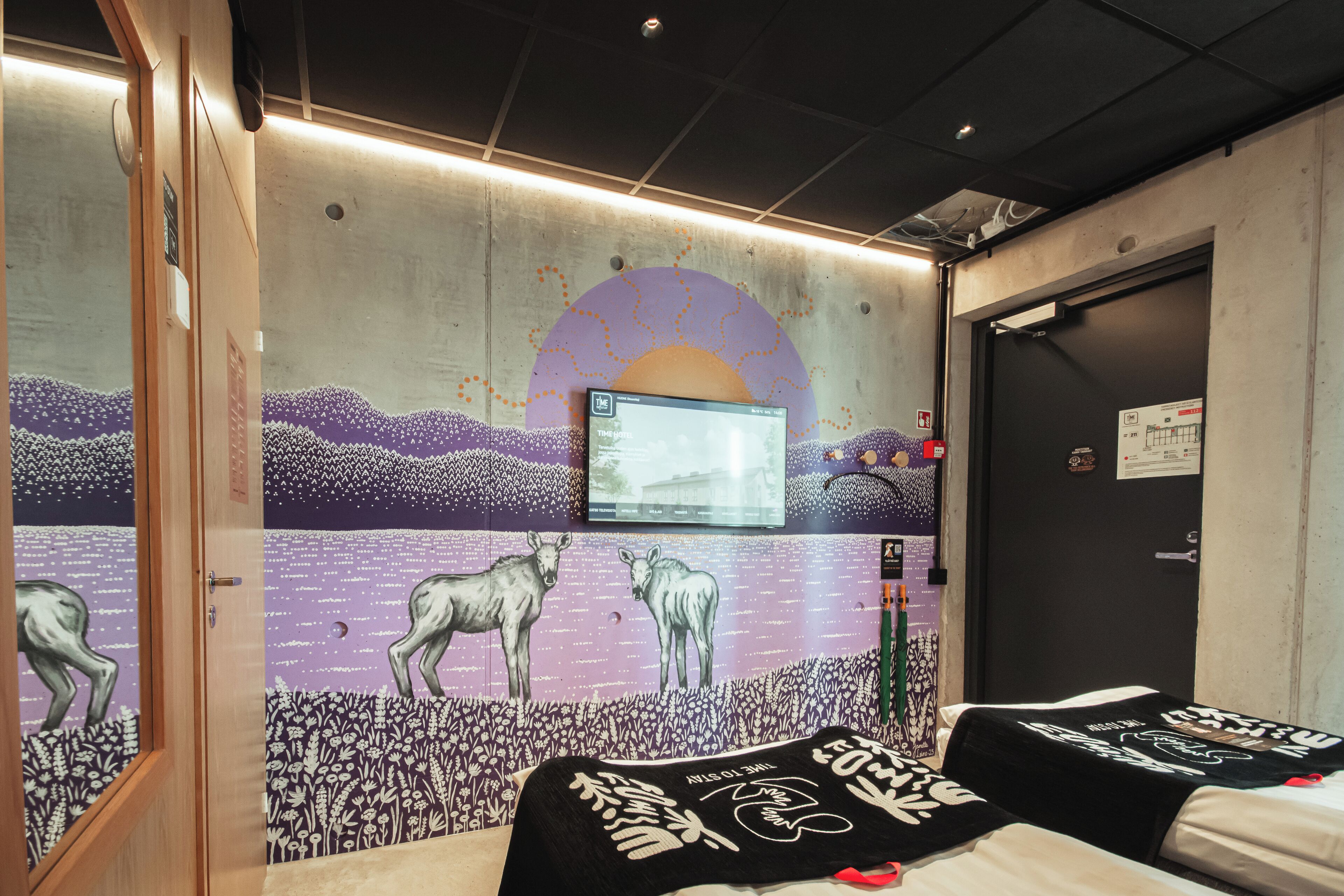 Una habitación con un mural de una puesta de sol y animales, una cama con una colcha de diseño blanco y negro, y un televisor en la pared.