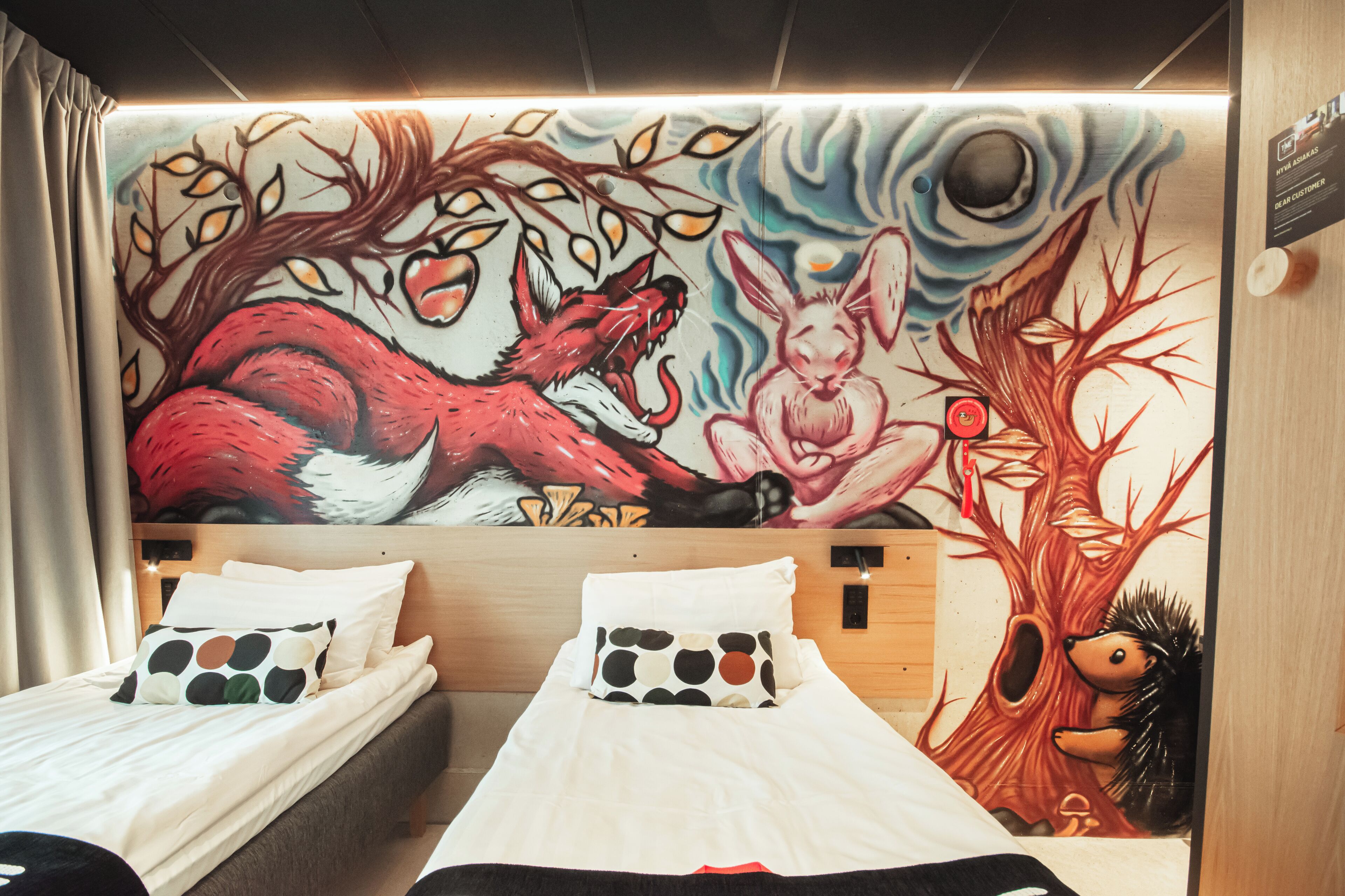 A hotel room with two beds, a mural with a wolf and a rabbit, and a tree with a hedgehog.