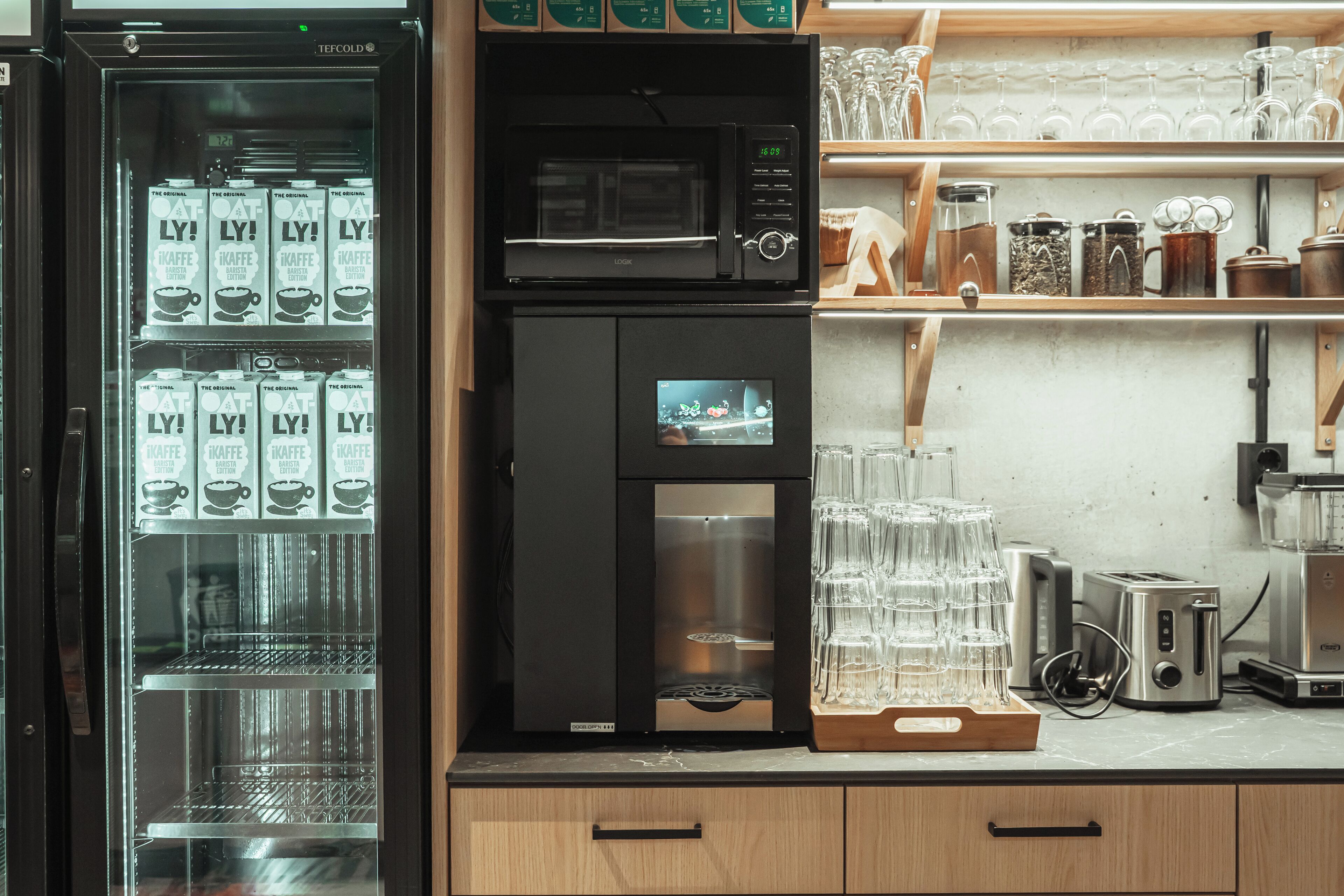 A kitchen area with a coffee machine, a microwave, and a refrigerator. There are also shelves with glassware and a coffee machine display.