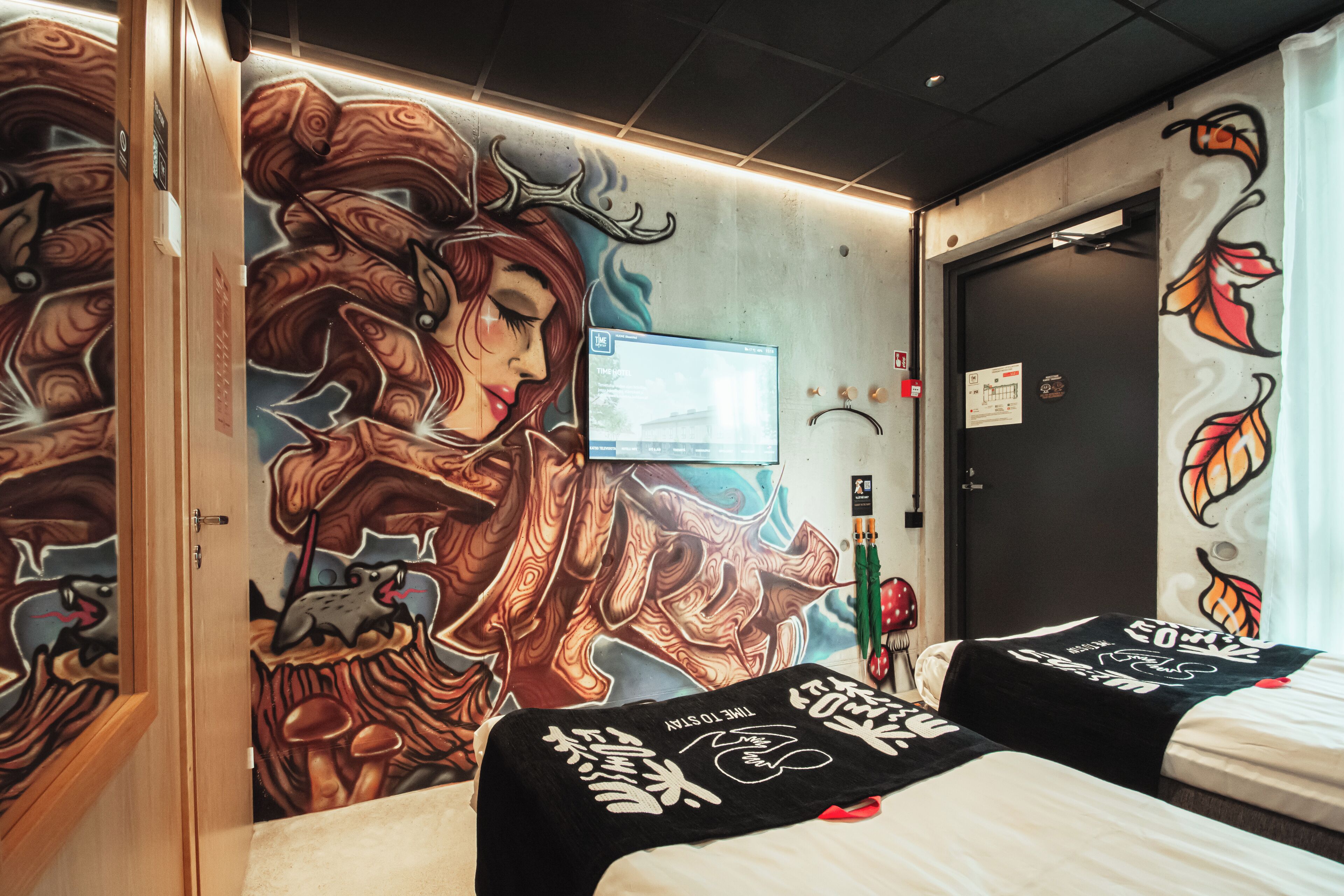 A hotel room with a mural on the wall, two beds with black and white bedding, a television, and a door with a sign.