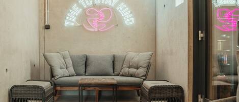 A cozy seating area with a sofa, two side tables, and wicker chairs. A neon sign on the wall reads "TIME TO STAY KIND."