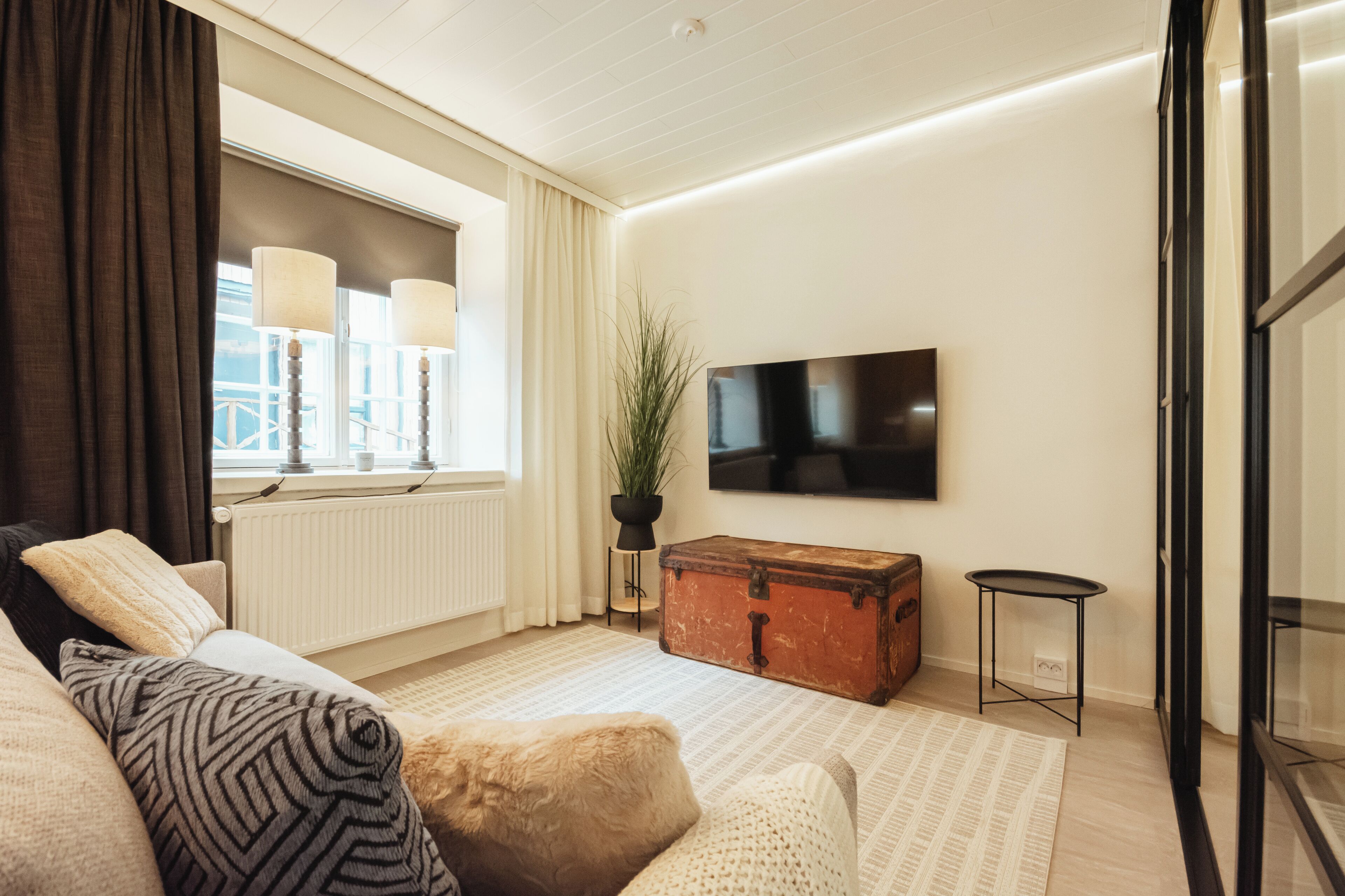 A modern living room with a sofa, a wooden chest, a potted plant, and a flat-screen TV mounted on the wall.