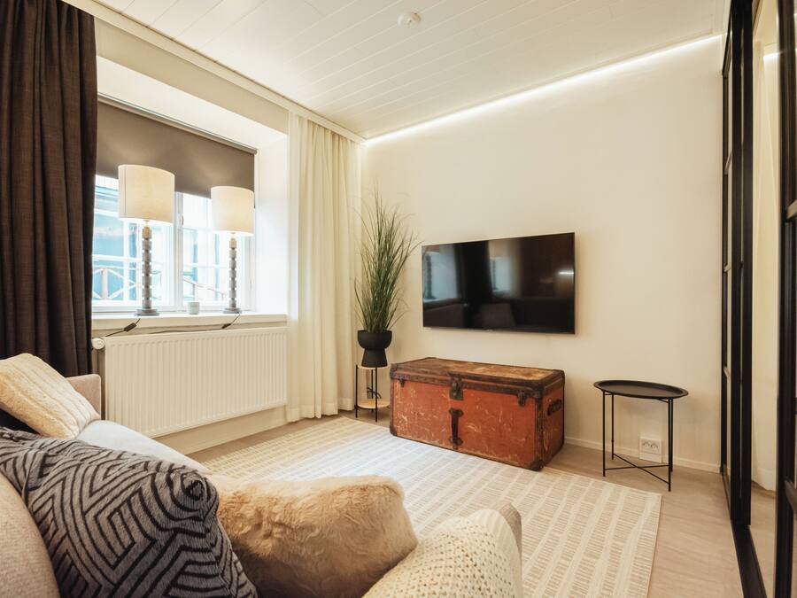 A modern living room with a sofa, a wooden chest, a potted plant, and a flat-screen TV mounted on the wall.