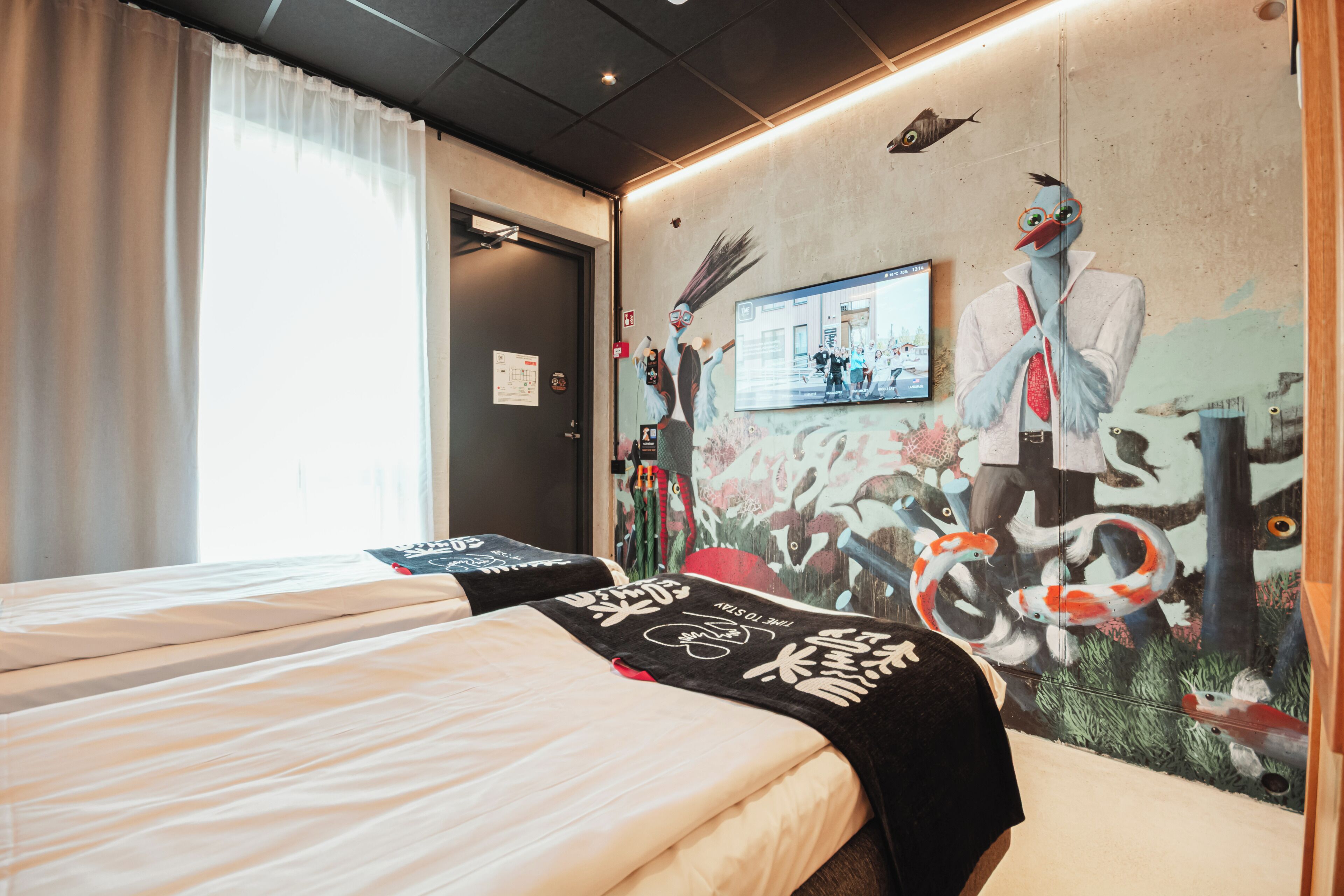 Una habitación de hotel con cama, un mural, televisor y ventana con cortinas.