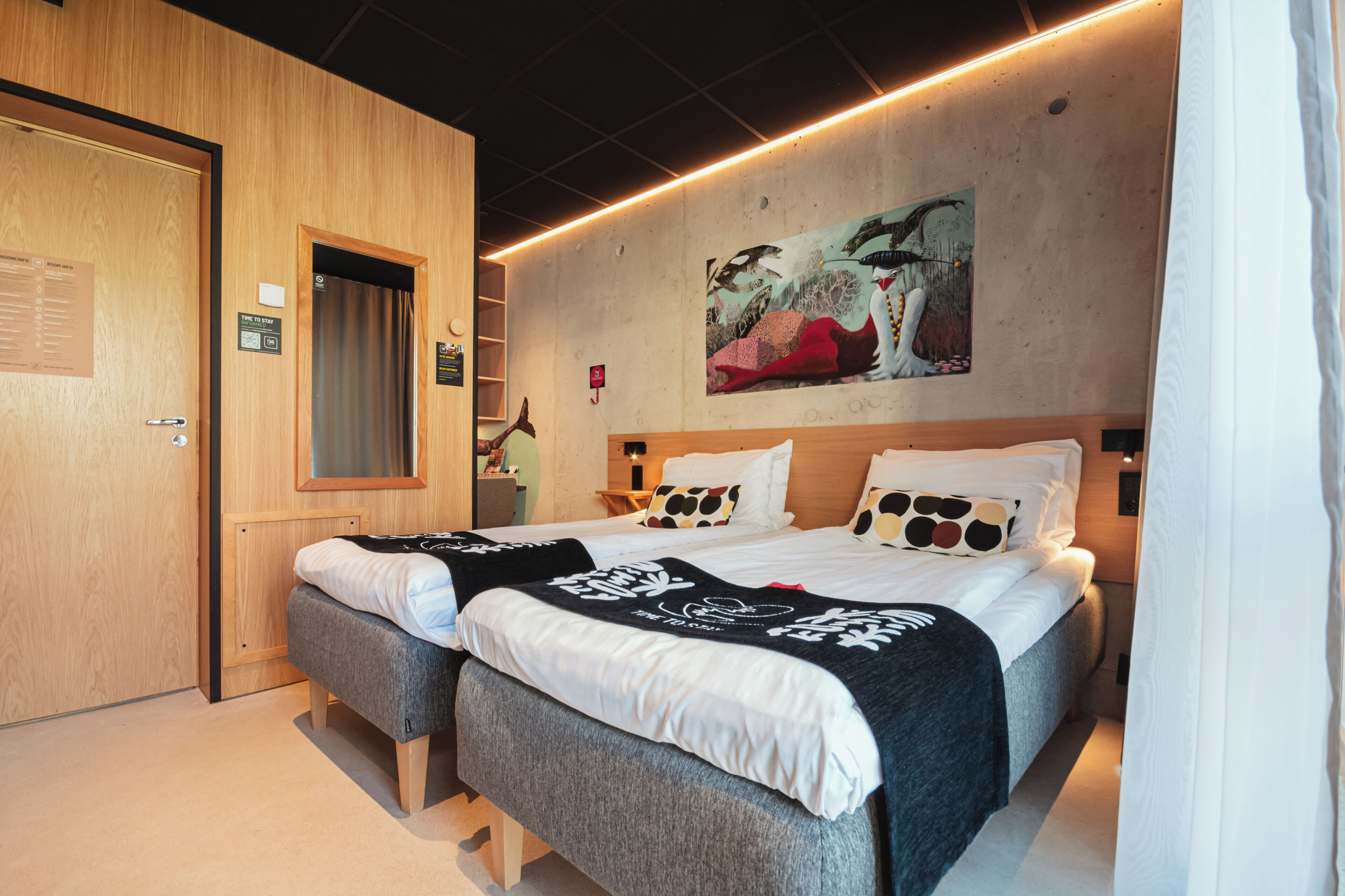 A hotel room with two beds, a wooden door, a painting on the wall, and a small desk with a lamp.