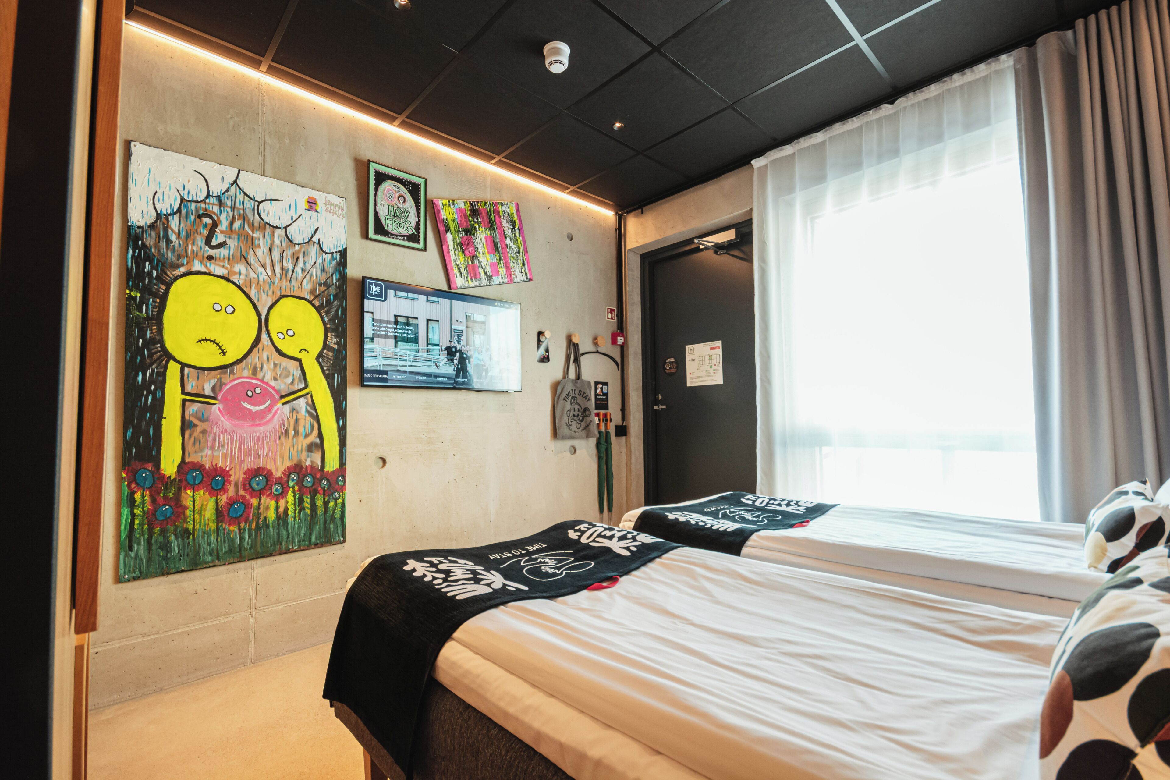 Ein Hotelzimmer mit zwei Betten, einem großen Fenster mit Vorhängen, einer Wand mit Kunstwerken und einer Tür.
