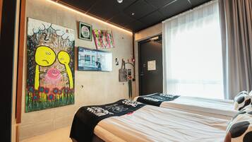 Ein Hotelzimmer mit zwei Betten, einem großen Fenster mit Vorhängen, einer Wand mit Kunstwerken und einer Tür.