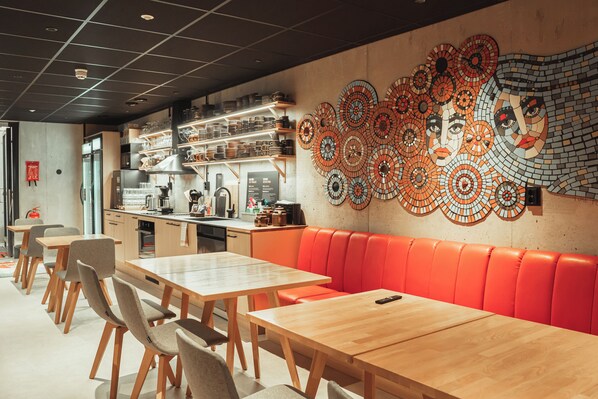 Un restaurant avec des tables en bois et des banquettes orange, une cuisine à l’arrière-plan et un mur décoré d’une mosaïque.