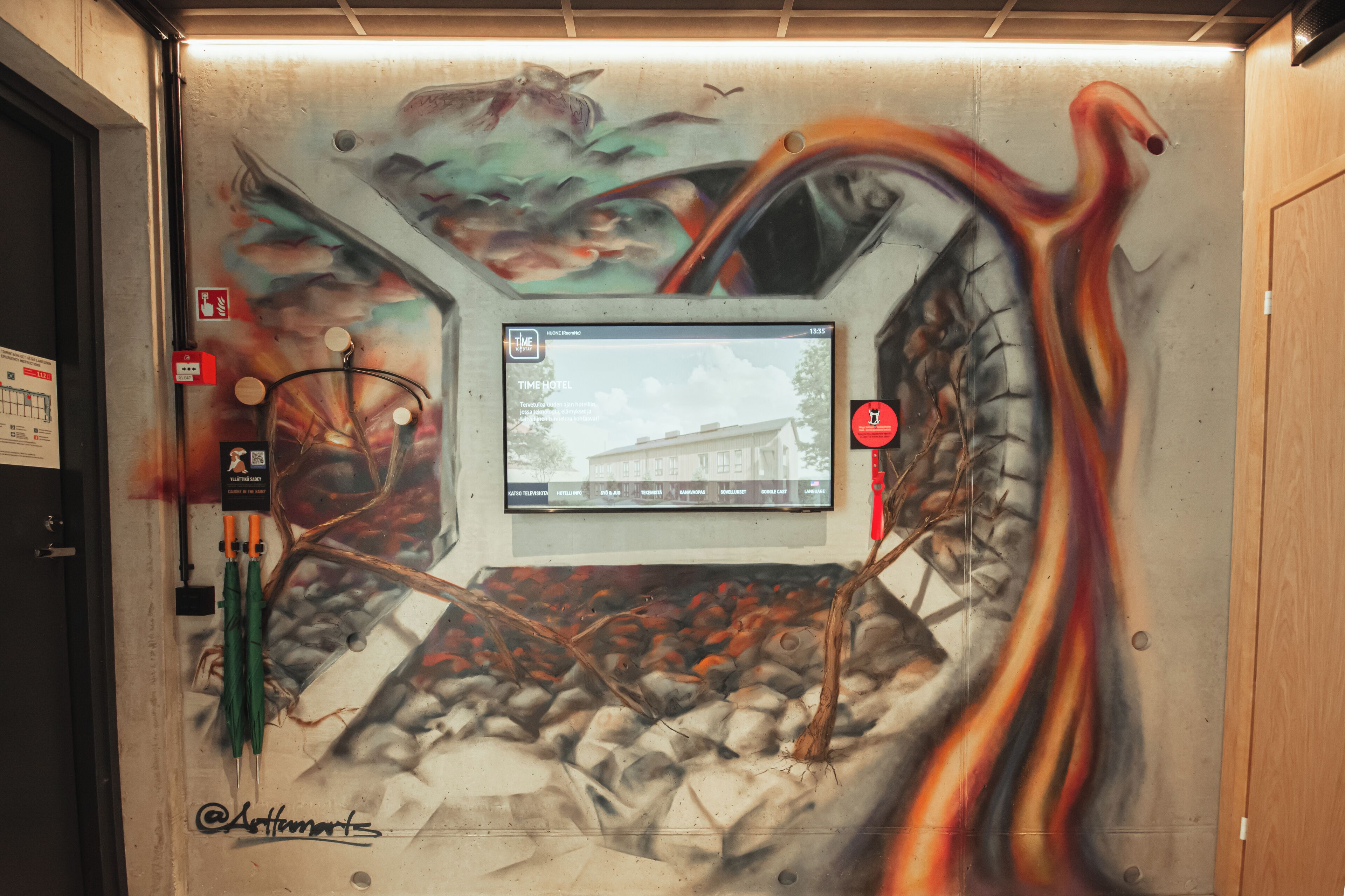 Pared con un mural de un coche y formas abstractas, una pantalla digital mostrando una imagen de hotel, y un dispositivo de activación de alarma contra incendios.