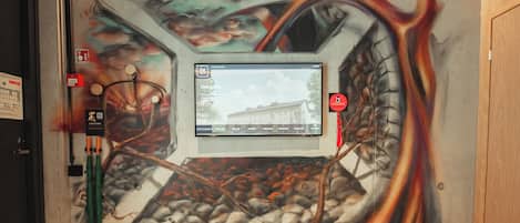 Pared con un mural de un coche y formas abstractas, una pantalla digital mostrando una imagen de hotel, y un dispositivo de activación de alarma contra incendios.