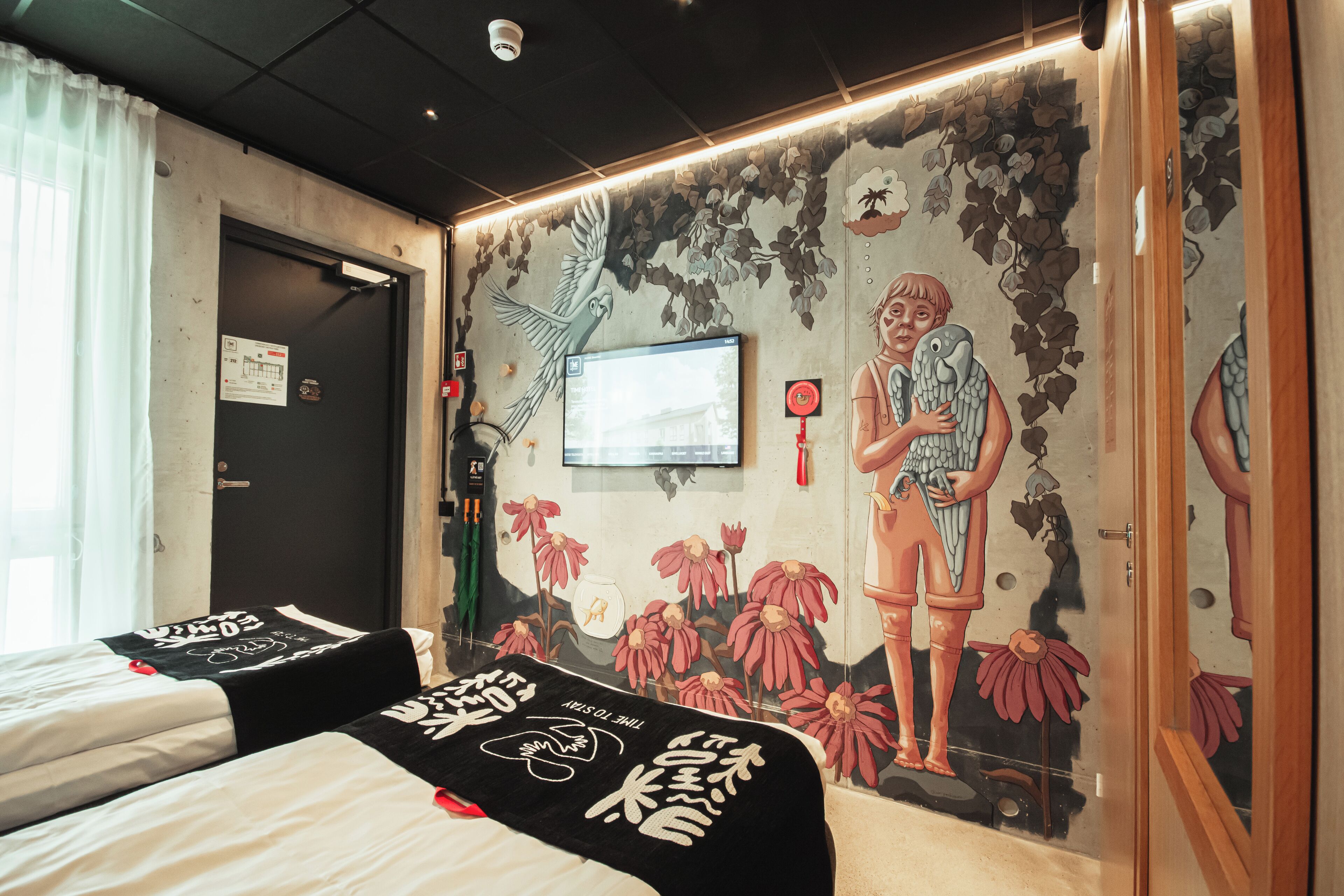 Habitación de hotel con dos camas, un mural con un pájaro y flores, un televisor y una puerta con un cartel.
