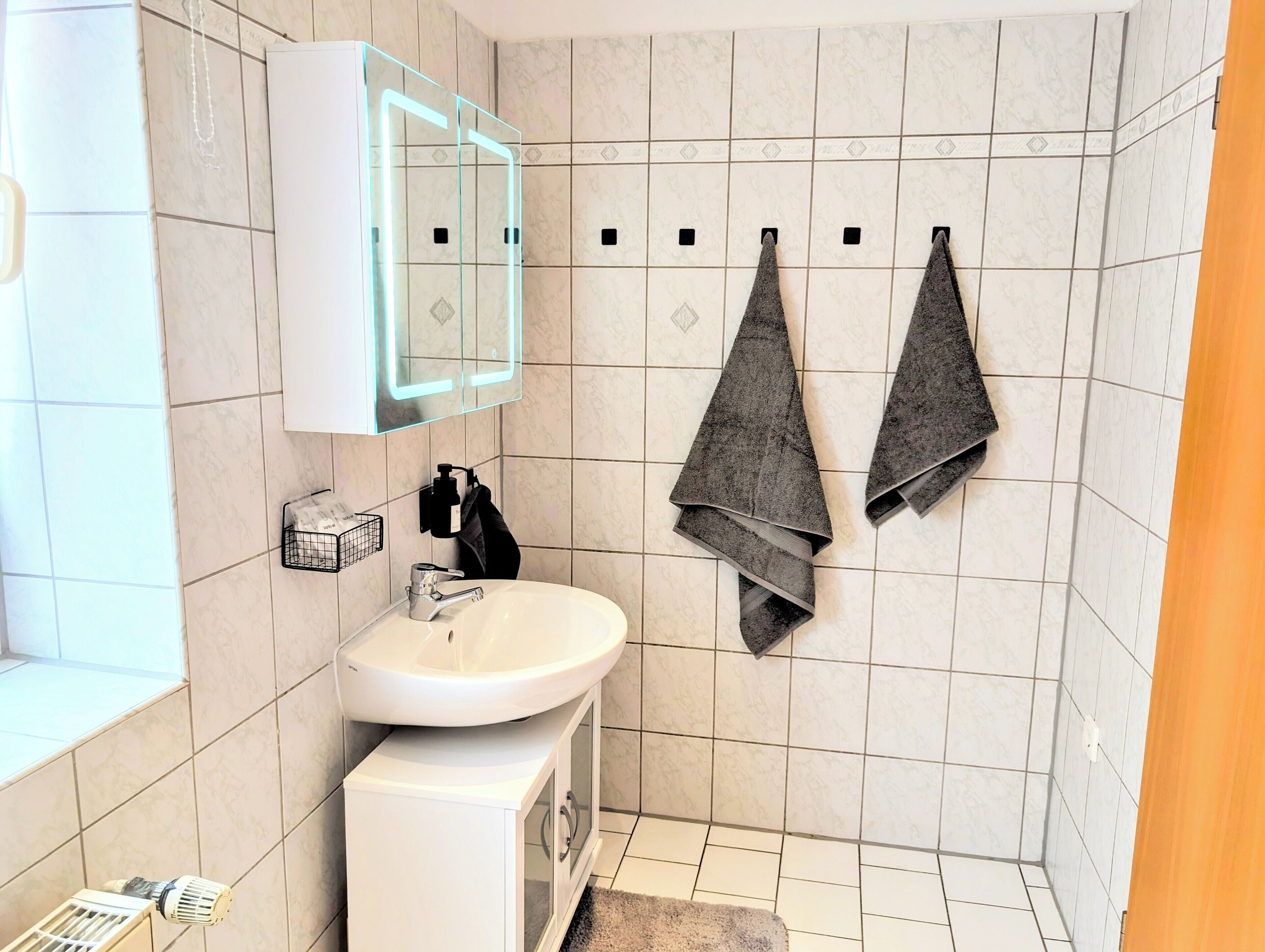 A bathroom with a white sink, a mirror with a light, and two towels hanging on hooks.