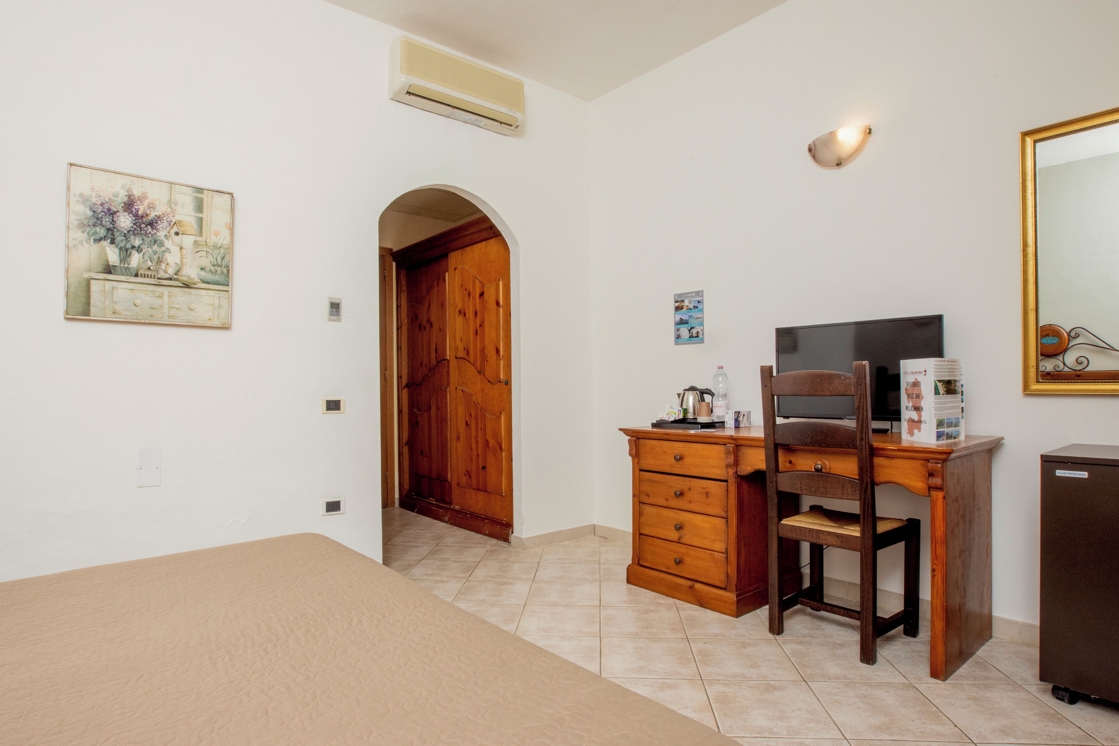 Un dormitorio con una cama, un escritorio con silla, un aparador, un espejo y una puerta que da a otra habitación.