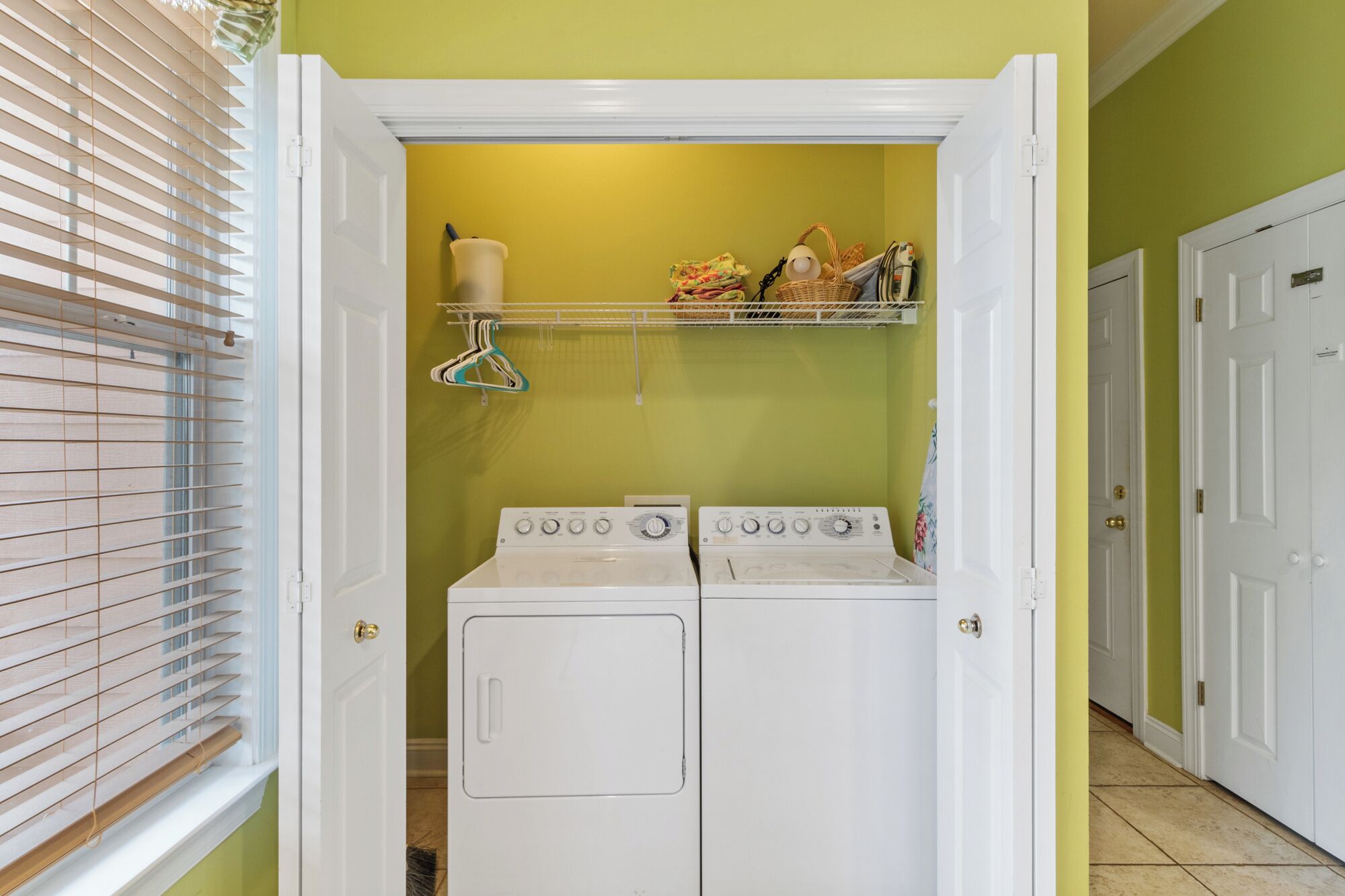 A laundry room with two white front-loading washing machines, a drying rack, and a shelf with laundry items.