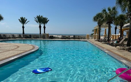 A clear blue pool with lounge chairs and palm trees, overlooking a beach.