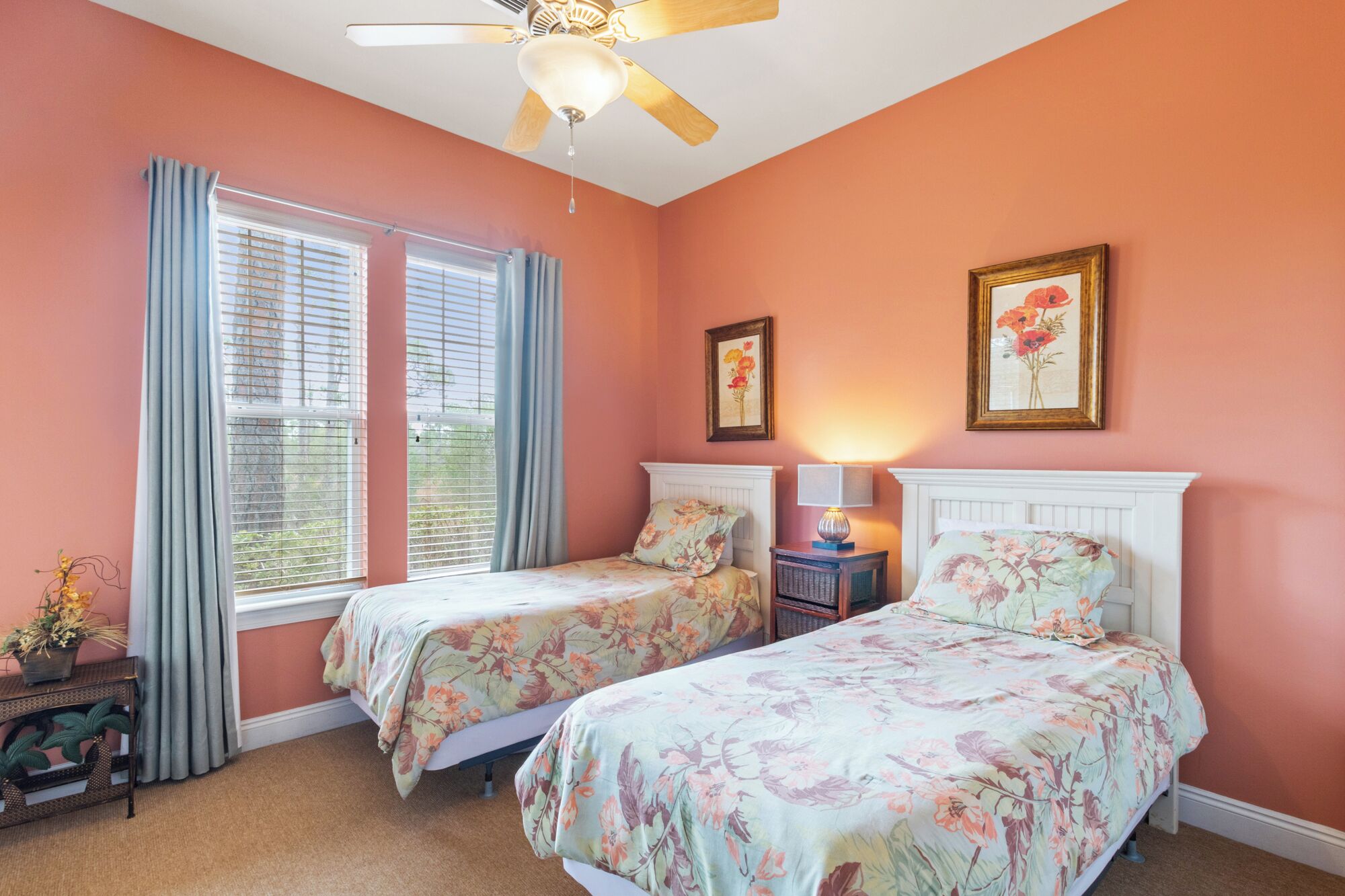 A bedroom with two beds, a ceiling fan, a window with curtains, and two framed floral paintings on the wall.