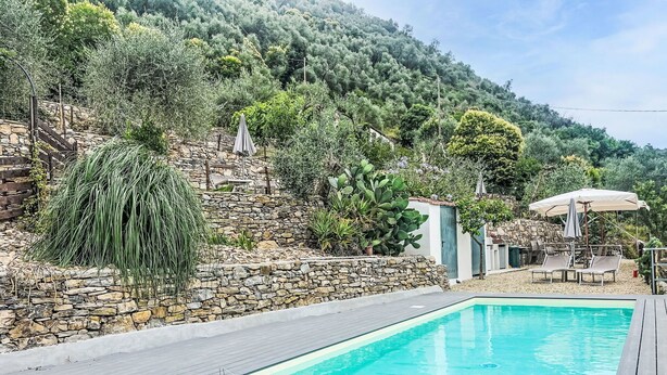 A swimming pool with a stone wall and wooden deck, surrounded by lush greenery and a hillside. - Cosy apartment for 4 people with WIFI, pool, TV, patio and pets allowed (Dolcedo)