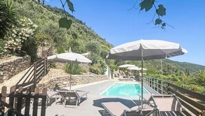 A poolside area with lounge chairs and umbrellas, a wooden deck, and a stone wall. - Cosy apartment for 4 people with WIFI, pool, TV, patio and pets allowed (Dolcedo)