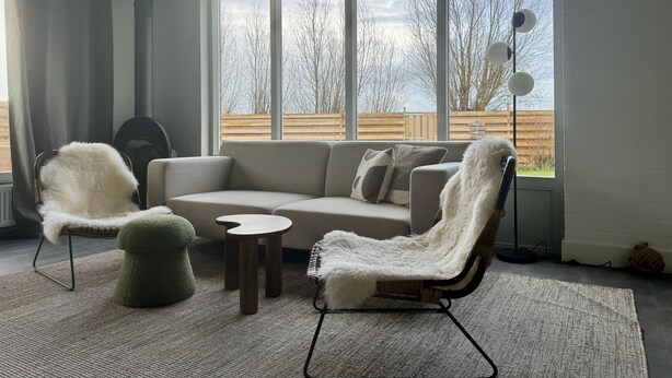 A modern living room with a sectional sofa, two armchairs, a small round table, and a large window with a view of trees and a wooden fence.