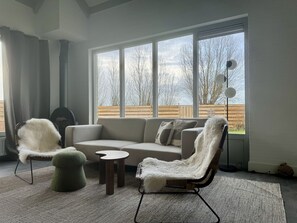 Un salon moderne avec un canapé d’angle, deux fauteuils, une petite table ronde et une grande fenêtre donnant sur des arbres et une clôture en bois.