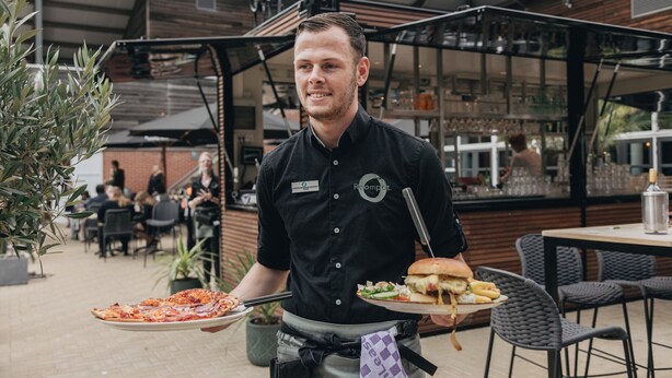 Henkilö, jolla on tarjotin, jossa on pizza ja hampurilainen, istuu ulkona ruokailualueella, jossa on pöytiä ja tuoleja.