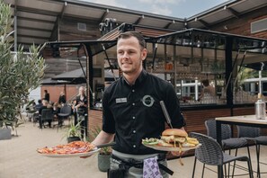 Una persona sosteniendo una bandeja con una pizza y una hamburguesa en un área de comedor al aire libre con mesas y sillas.