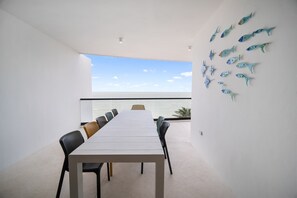 A dining area with a long table and chairs, a balcony with a view of the sea, and decorative fish wall art.