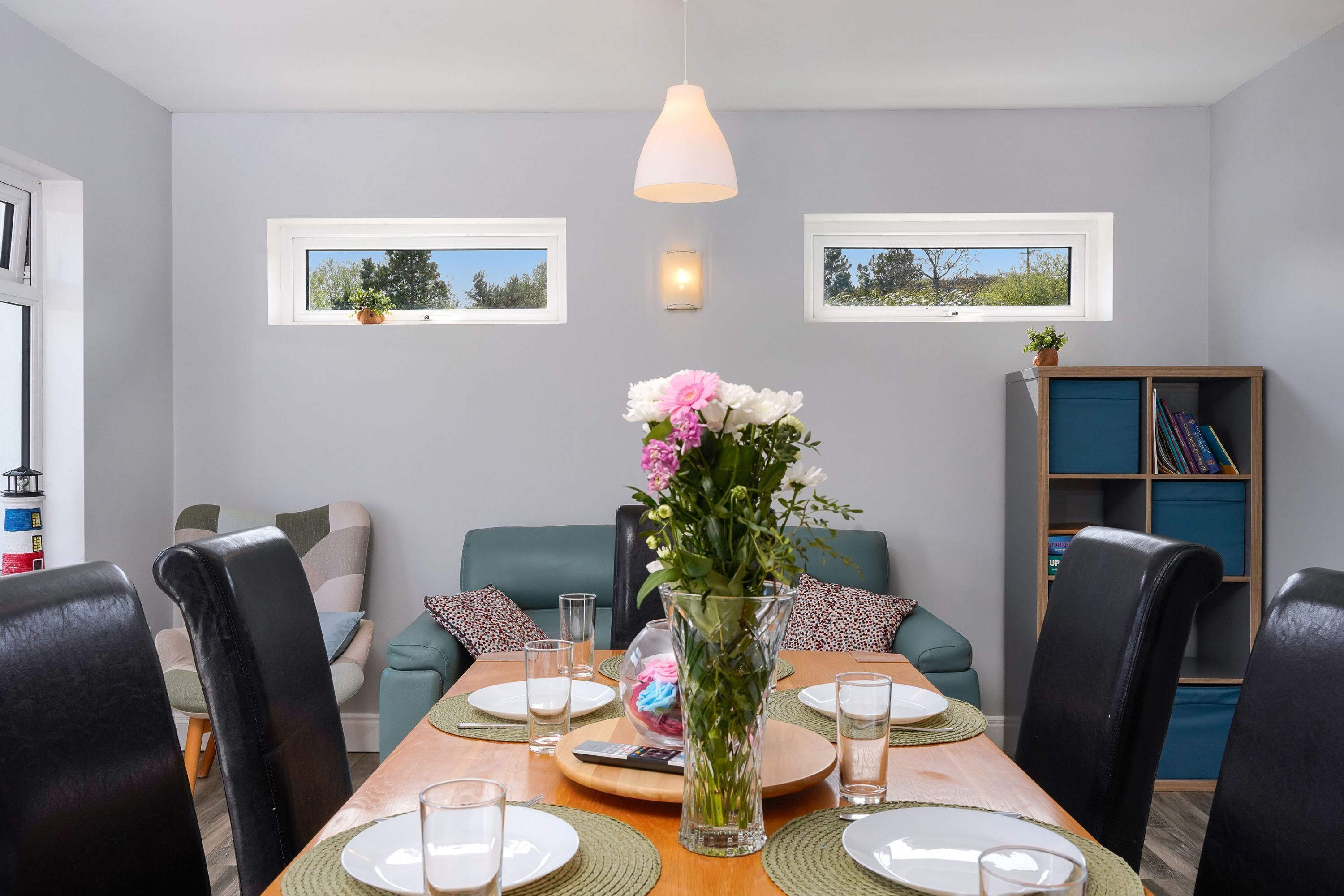 En matplats med ett träbord dukat för fyra personer, omgivet av svarta stolar. Det finns två fönster med utsikt över grönskan, en bokhylla med böcker och en vas med blommor på bordet.