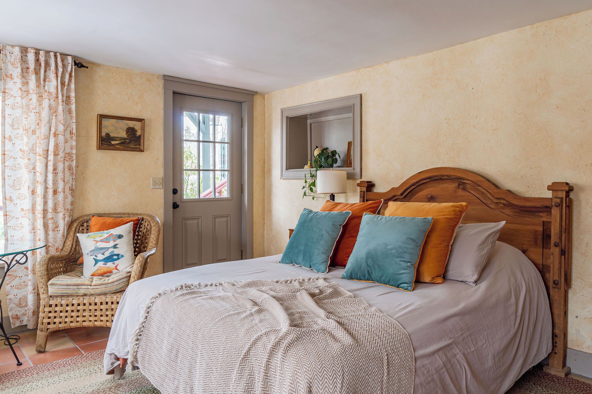 Un dormitorio con una cama de madera, una silla de mimbre, una puerta y un espejo colgado en la pared.