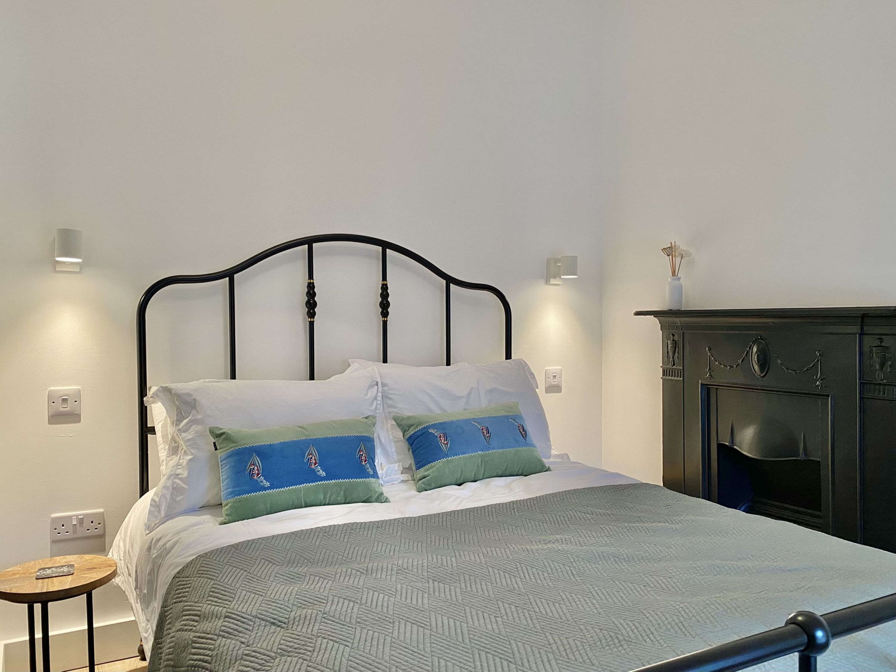A neatly made bed with a patterned bedspread and decorative pillows, a small round table beside it, and a dark wooden cabinet against the wall.