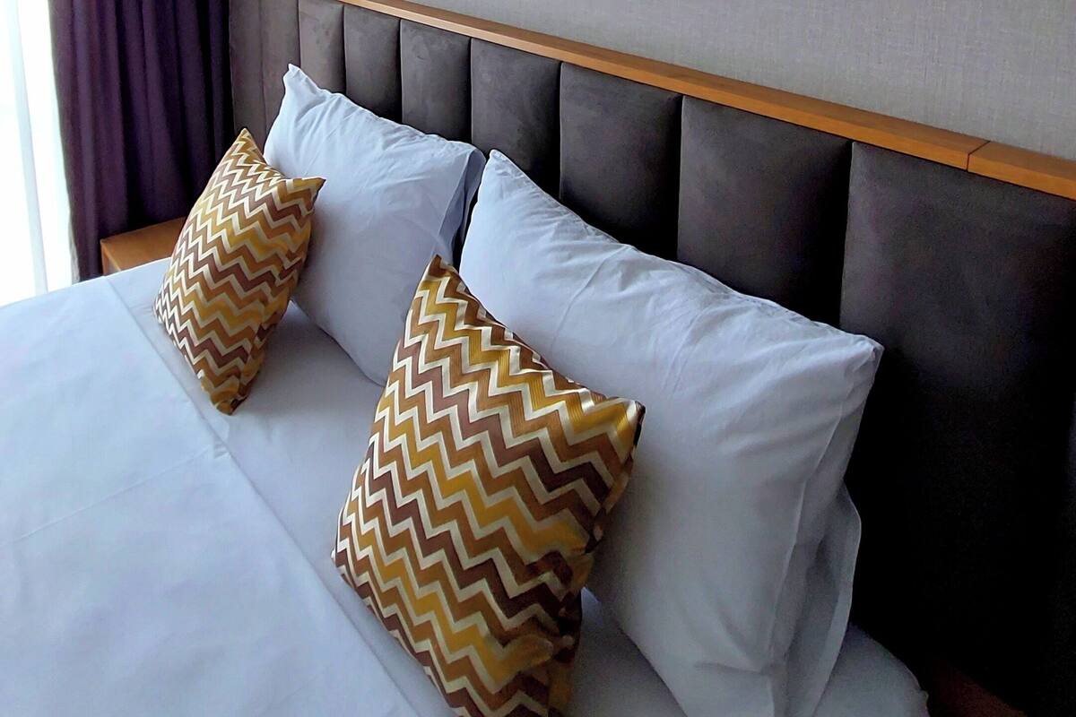 Una habitación de hotel con cama, dos almohadas, una obra de arte enmarco y una lámpara fijada a la pared.