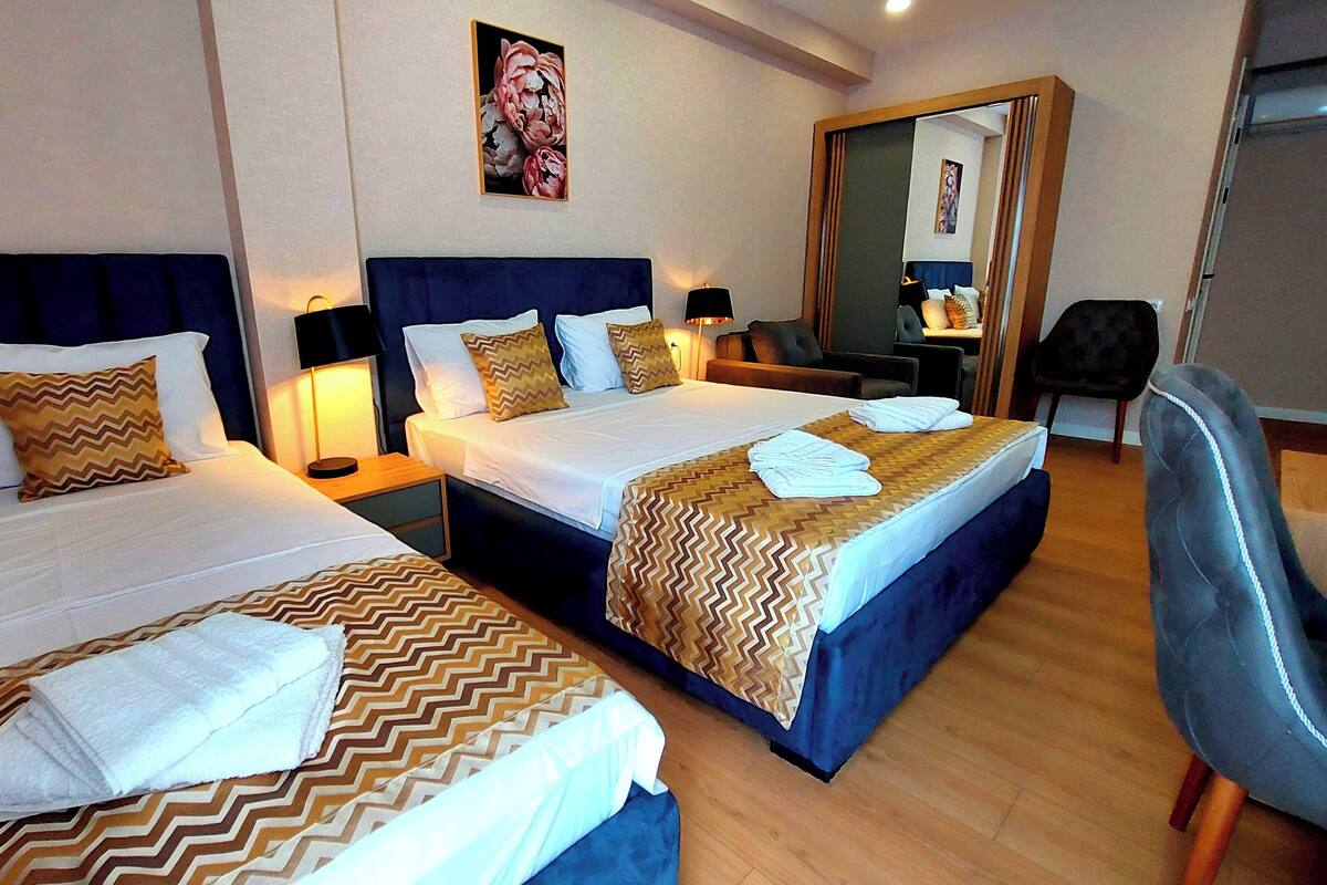 Habitación de hotel con dos camas, cada una con cubrecama estampado, una mesita de noche con lámpara, una silla y un espejo en la pared.