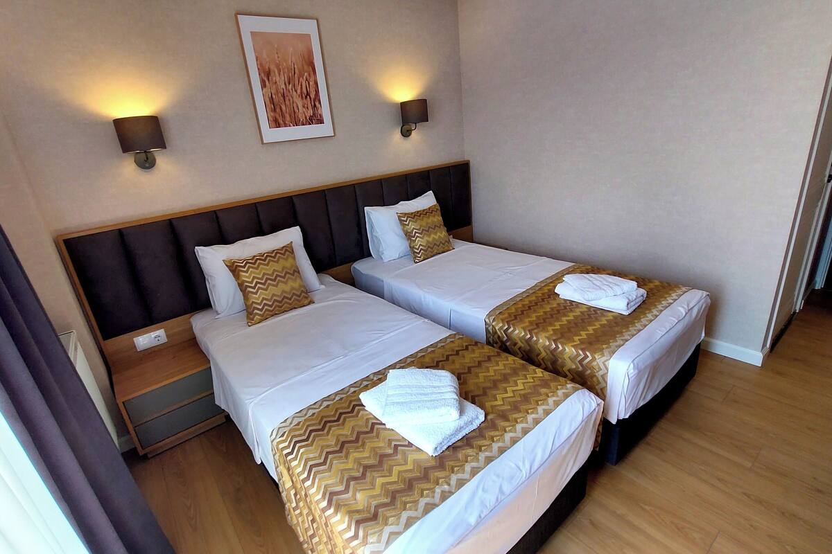 Una habitación de hotel con dos camas, cada una con cubrecama dorado y blanco, cabecero de madera y mesita de noche con lámpara.