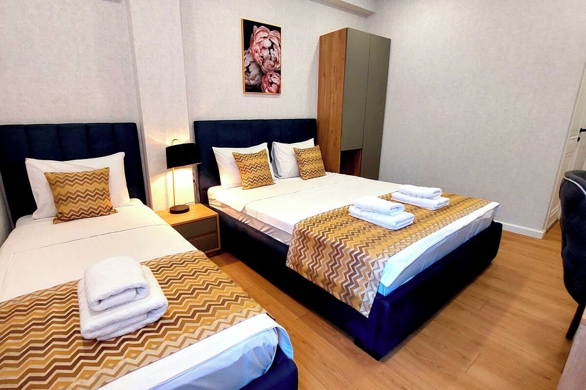 Habitación de hotel con dos camas, cada una con colcha estampada, almohadas y toallas dobladas. Hay una mesita de noche con lámpara, un armario y un cuadro en la pared.