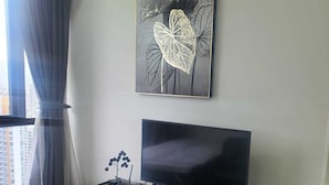 En moderne stue med et stort fladskærms-tv på et træpanel, kunst på væggen af abstrakte blade, og udsigt til bylandskab gennem et vindue med gardiner.