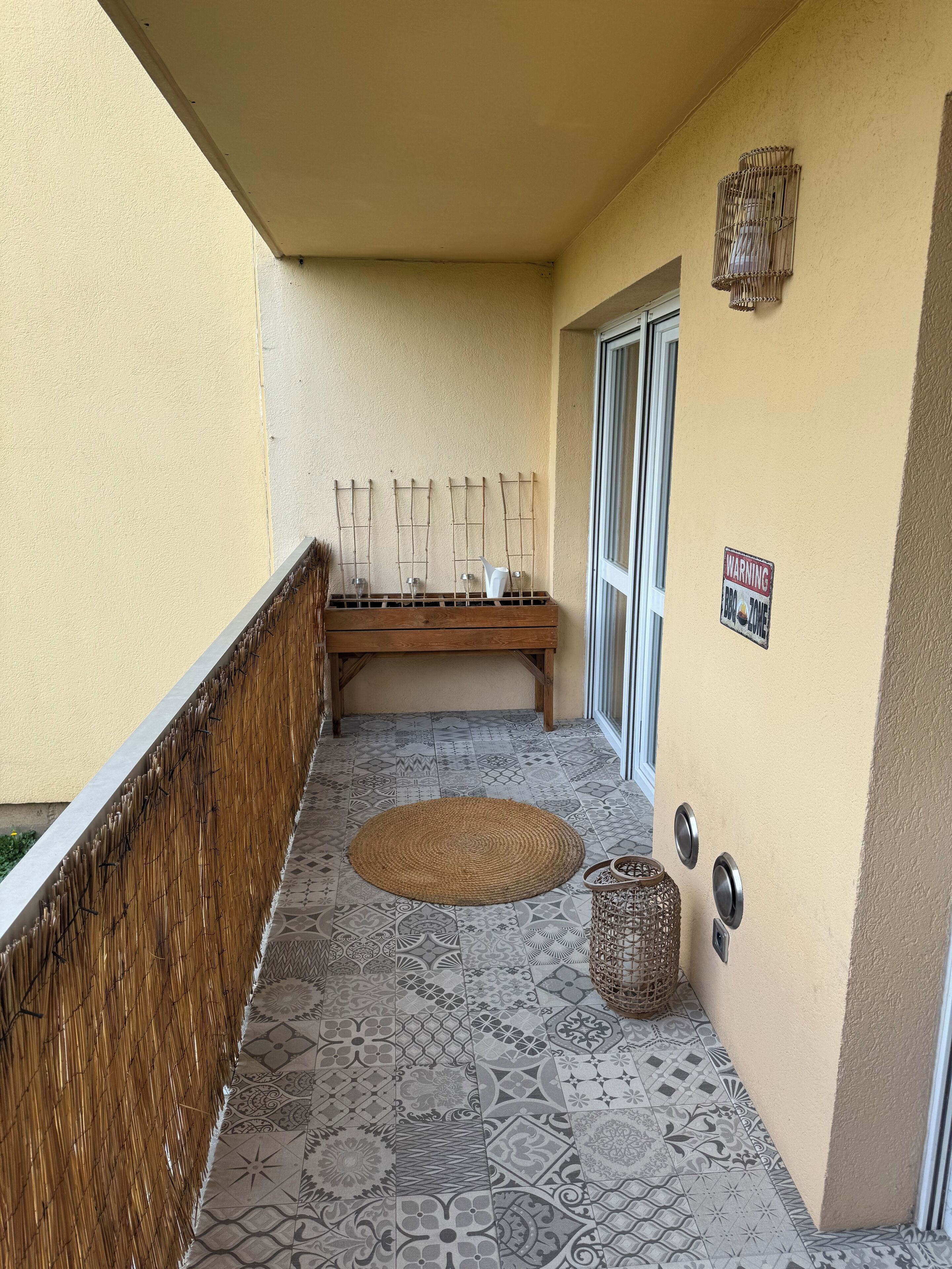 Een balkon met een patroon in de vloer, een houten bankje, een geweven mat en een waarschuwingsbord.
