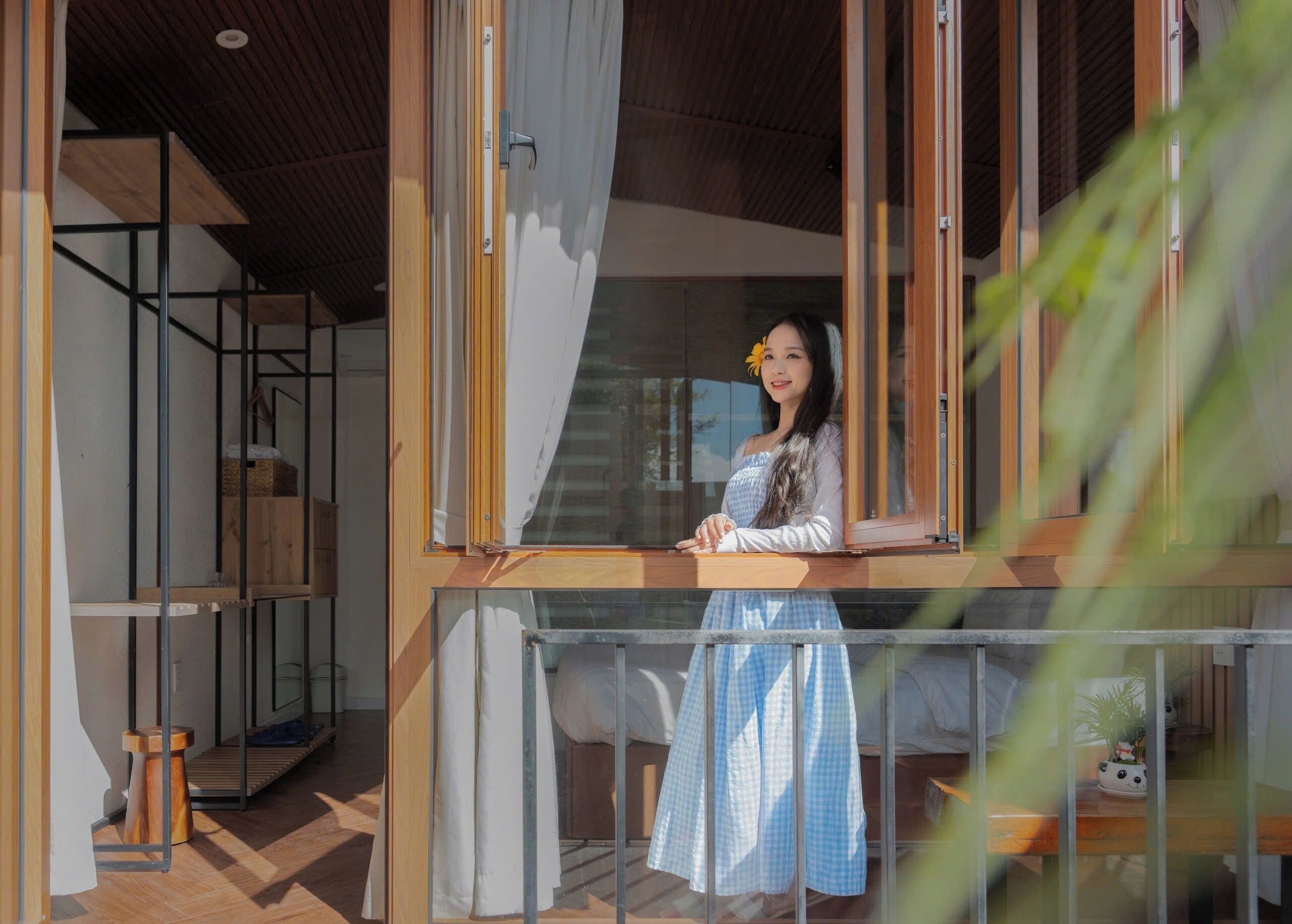 Una persona de pie en un balcón con barandilla de madera, una ventana con cortinas y vista al interior de una habitación con cama y planta.
