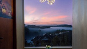 A room with a large window offering a view of a sunset over a landscape, two wooden stools, a small table with a vase, and a dog. - Mê Villa Dalat (Xuan Huong - Da Lat)