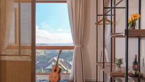 A modern living room with a large window, a guitar, a blue rug, and a wooden bookshelf. - Mê Villa Dalat (Xuan Huong - Da Lat)